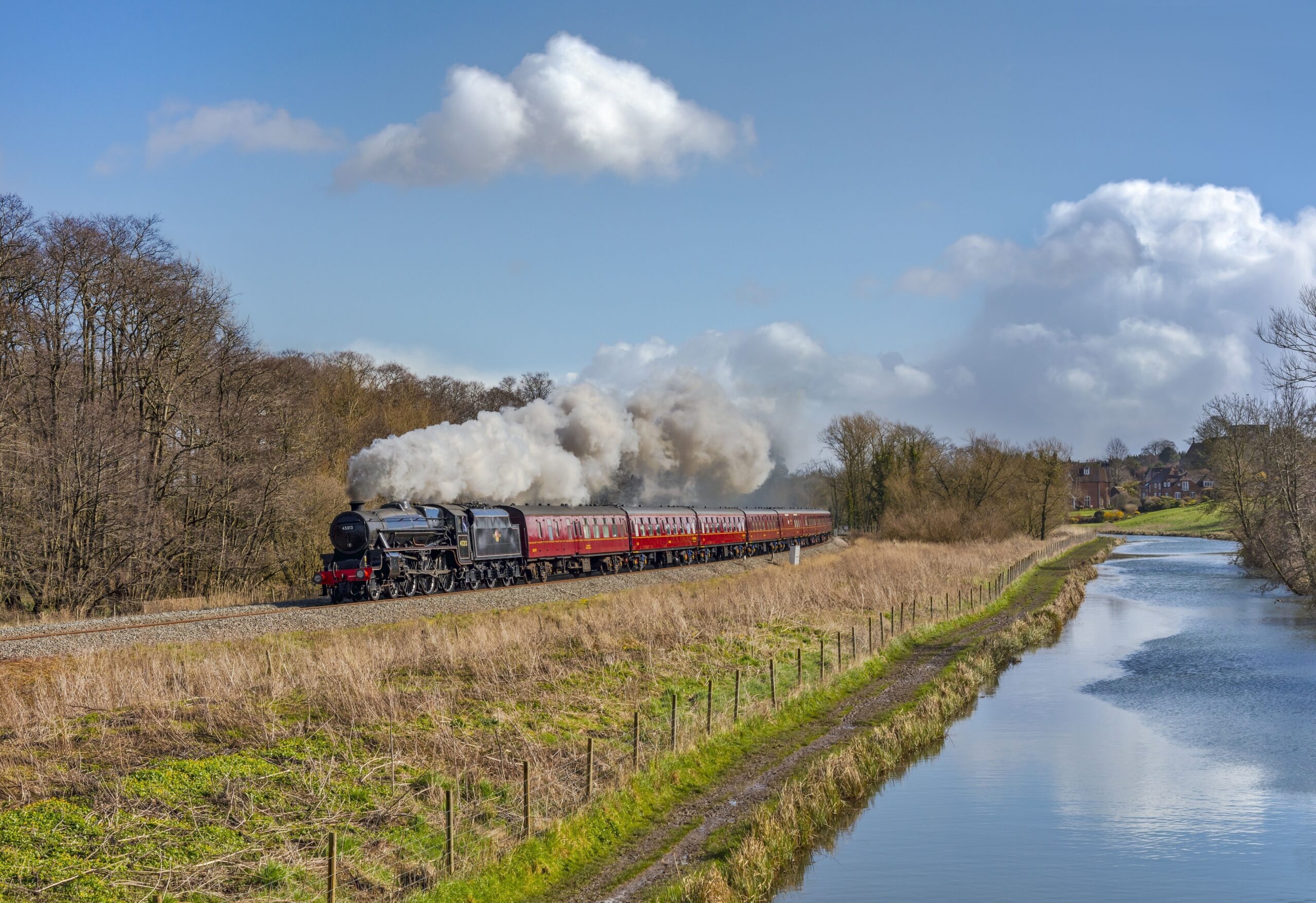 A steam train chugs along a picturesque countryside, leaving a trail of smoke against a clear blue sky.